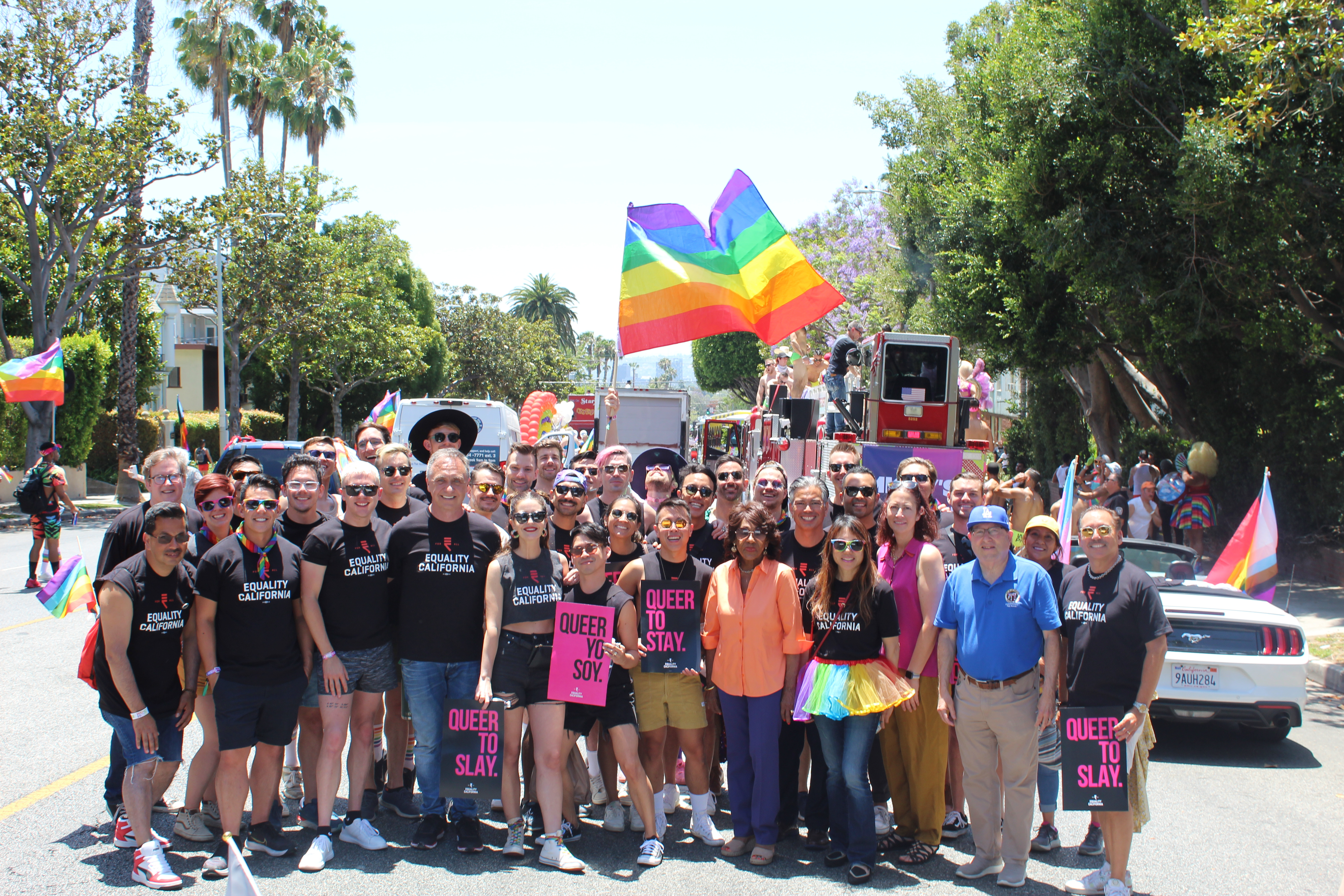 A group of people stand together holding a rainbow flag high, smiling at the camera.