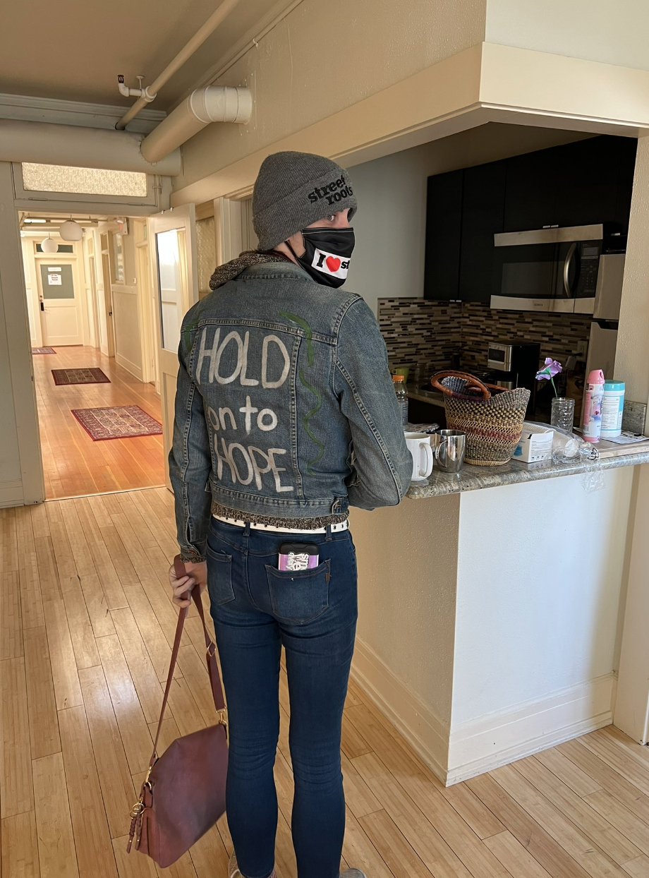 A person turned slightly away from the camera wears a mask, holds a maroon bag, and wears a grey jacket that says "hold onto hope"