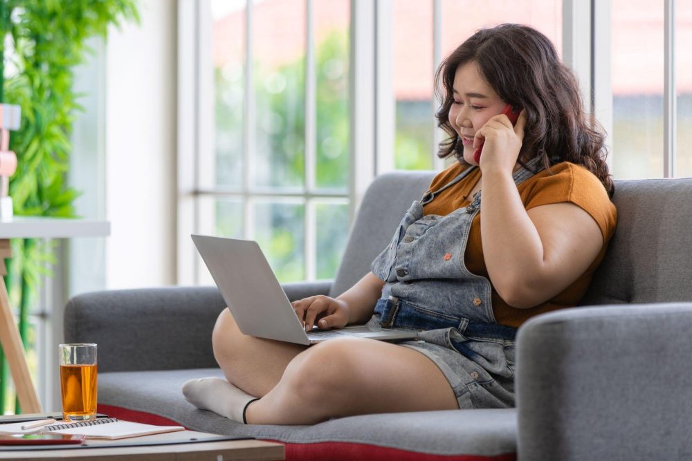 Woman wearing overalls sits on a couch while talking on the phone and using her computer