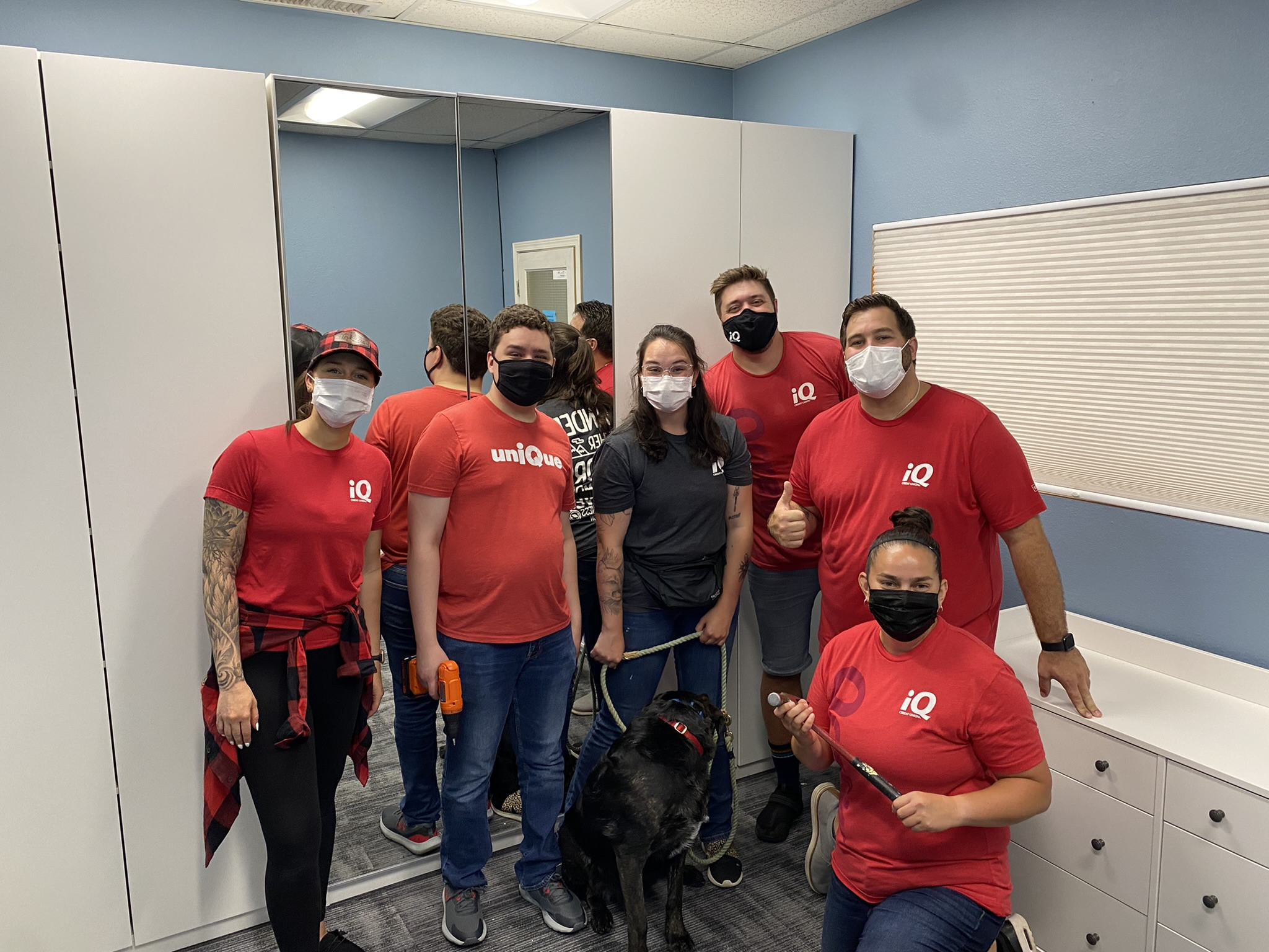 Six people in front of a tall white closet. Full-length mirrors are on the middle doors. They are all wearing masks, t-shirts, and either jeans or jean shorts. One person holds a medium-sized black dog on a leash. A person kneeling on the right side of the group holds a hammer.