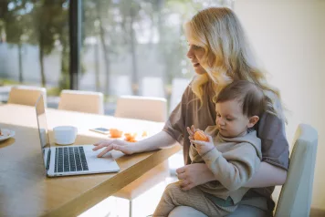 A woman sits at a table holding her child and using a laptop computer