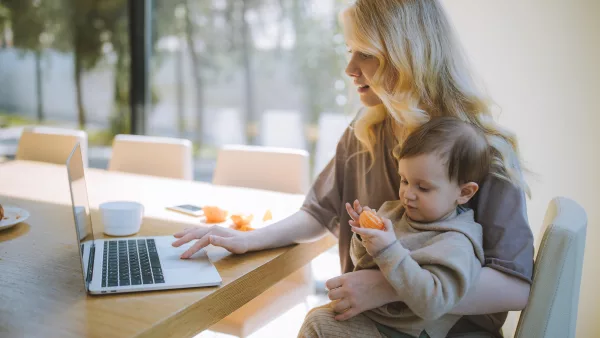 A woman sits at a table holding her child and using a laptop computer